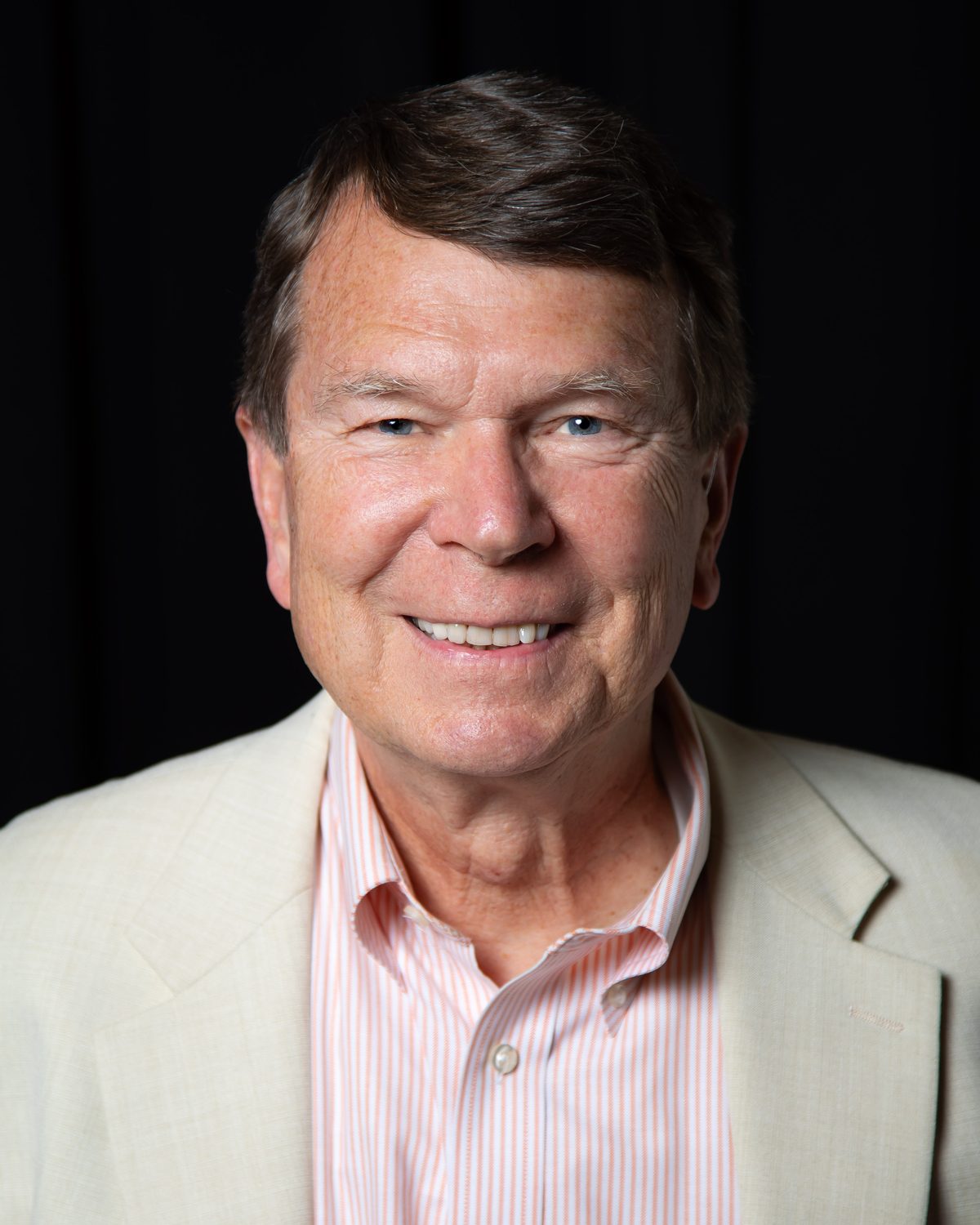 Steve Hauser in a cream jacket and striped shirt, smiling, against a black background
